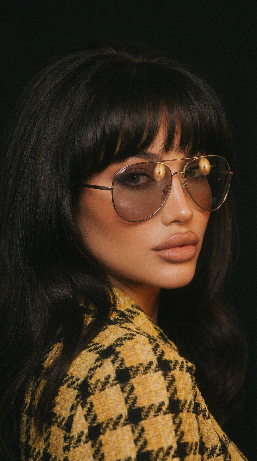 A high-fashion retro portrait showcasing a woman in oversized tinted aviator glasses, featuring a textured yellow outfit against a deep backdrop.