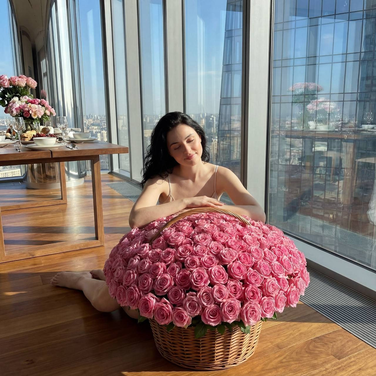 A girl sitting on the floor in a penthouse, surrounded by a massive floral bouquet of dense roses, smiling softly.