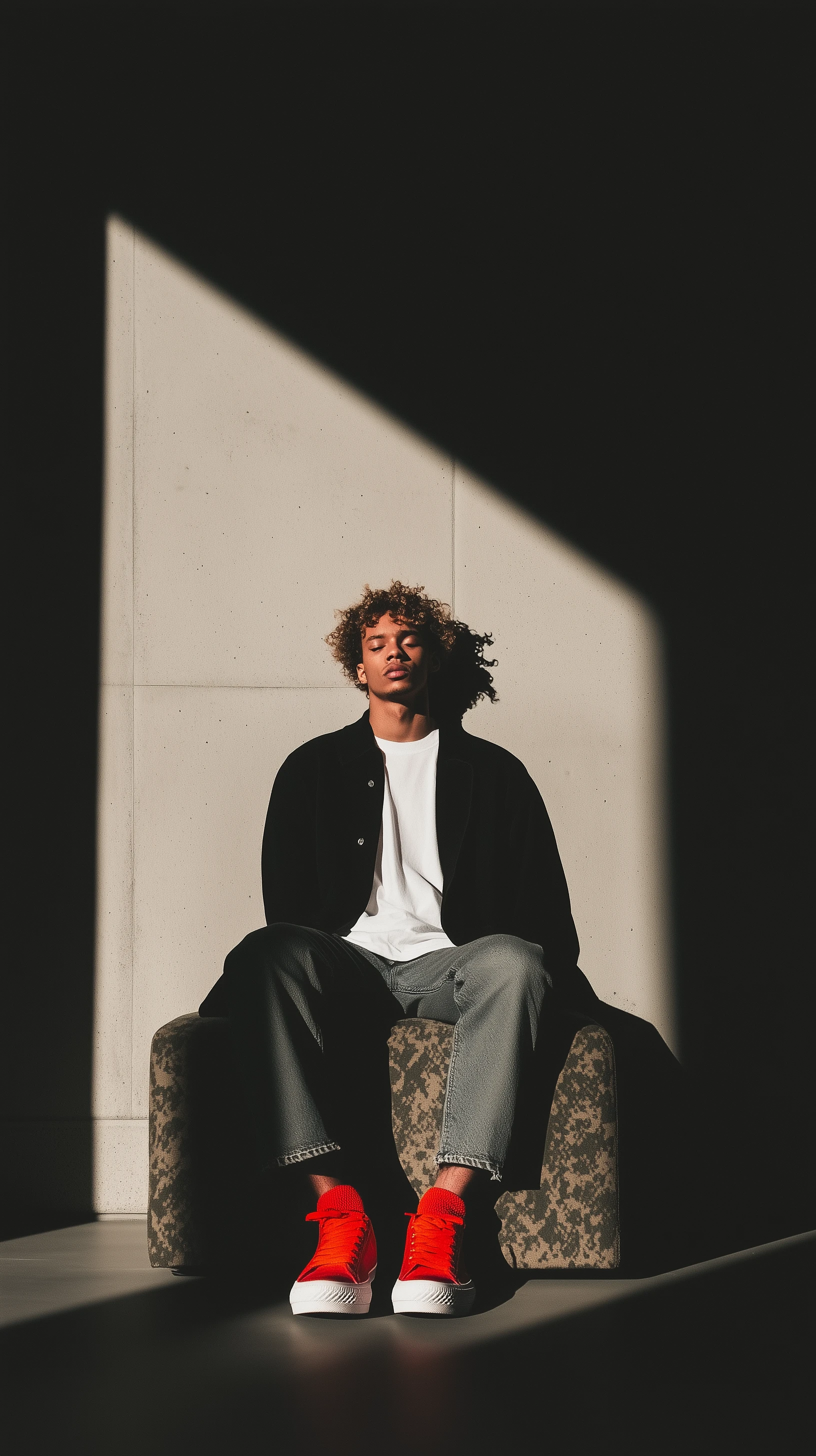 A male model lounging on a worn camouflage sofa, dressed in a white T-shirt and black cardigan, against a textured concrete wall.