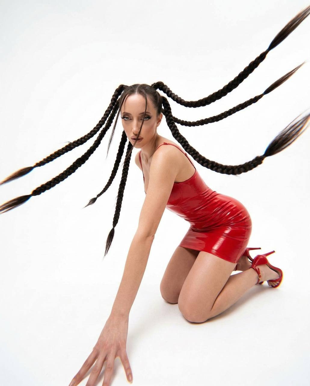 A woman in a red latex mini dress kneeling confidently on a white background, her long braids spread dynamically.