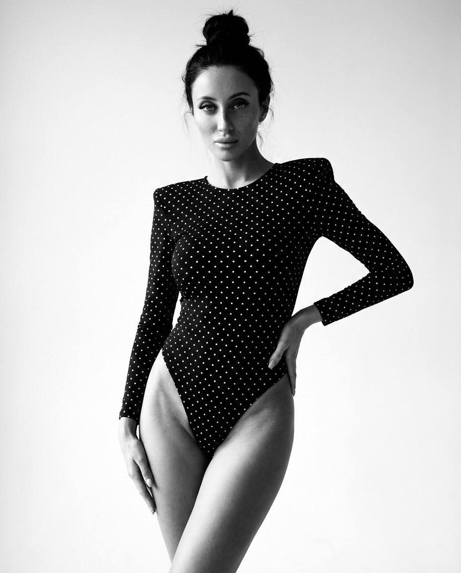 A slender model in a high-slit black bodysuit adorned with tiny polka dots, posed elegantly against a minimalist light background.