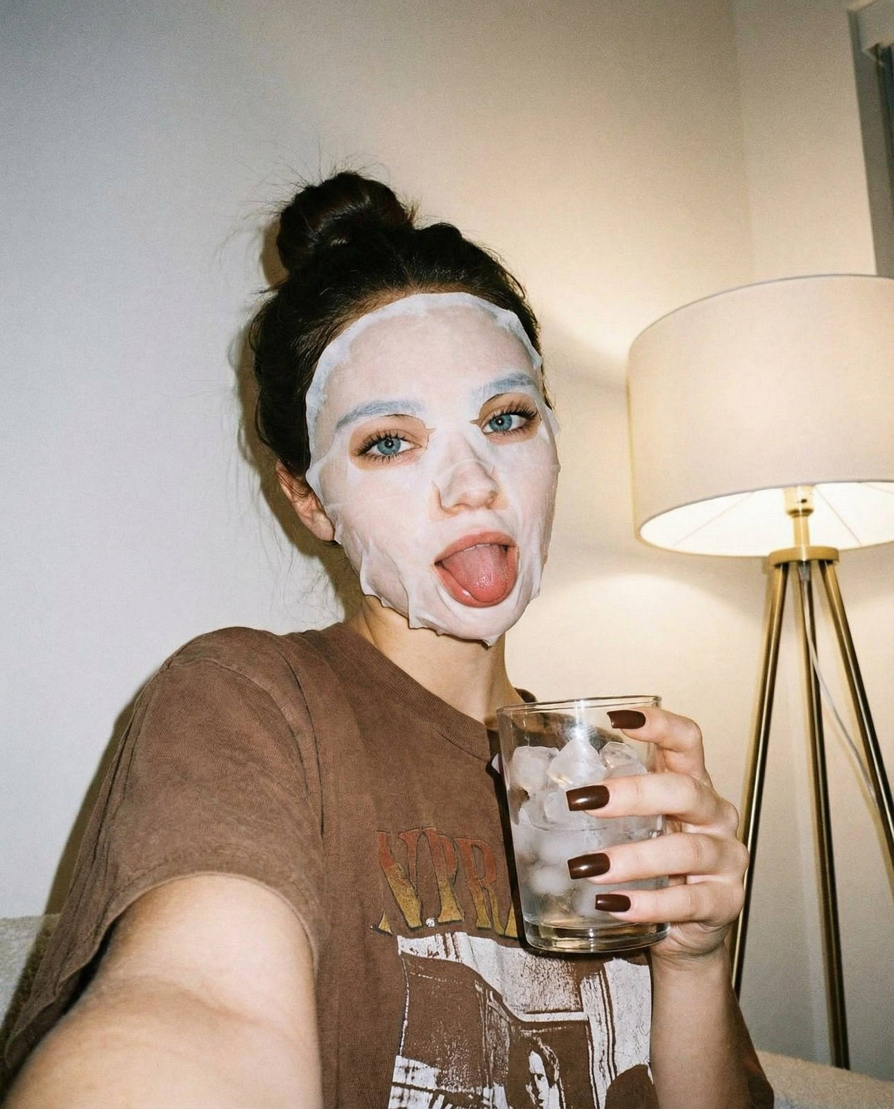 Portrait of a girl wearing a skincare face mask, playfully sticking out her tongue while holding a glass with ice cubes, in warm home lighting.