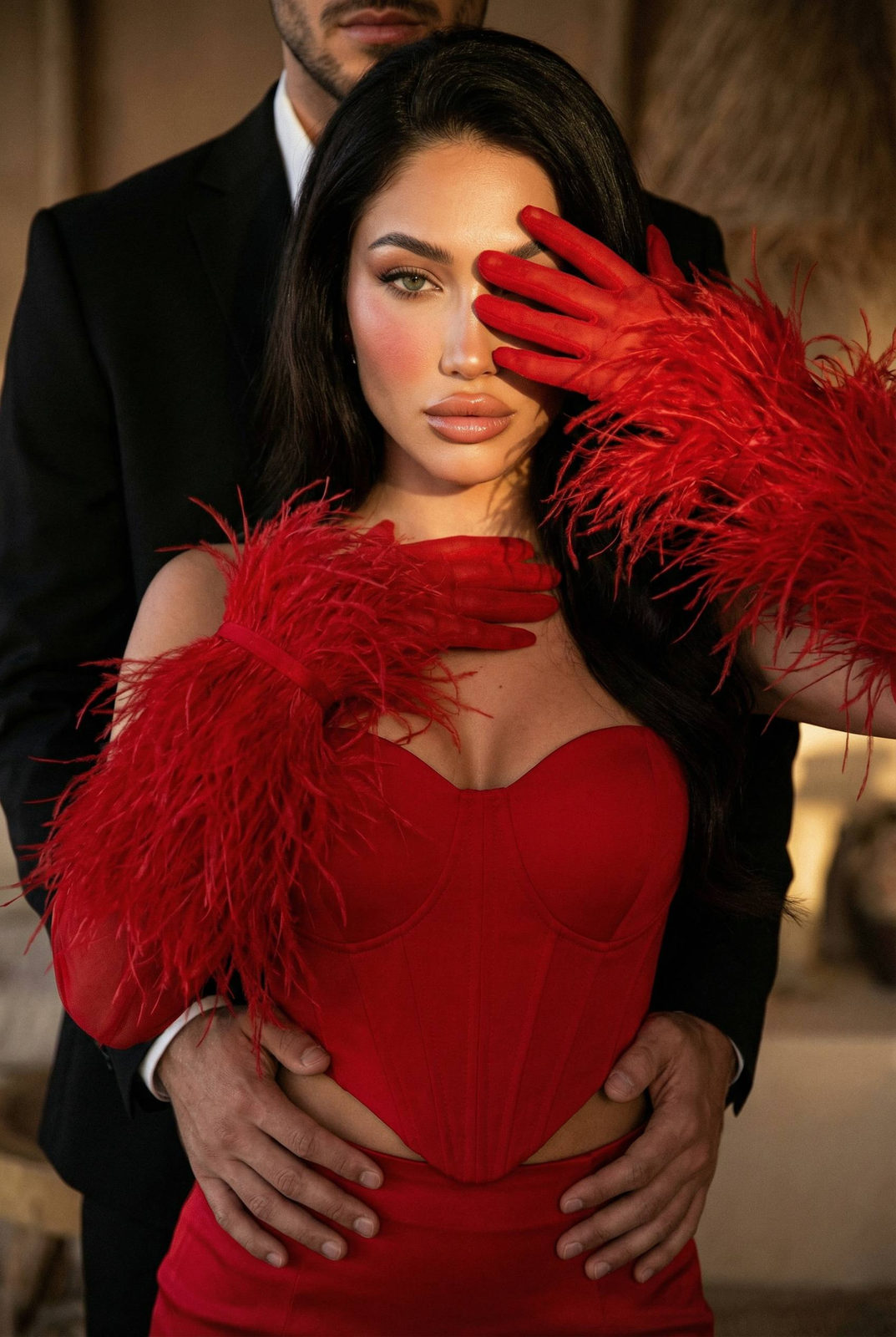 High-fashion portrait of a woman in a red corset with feather gloves, one eye visible, dramatic pose against neutral warm lighting.