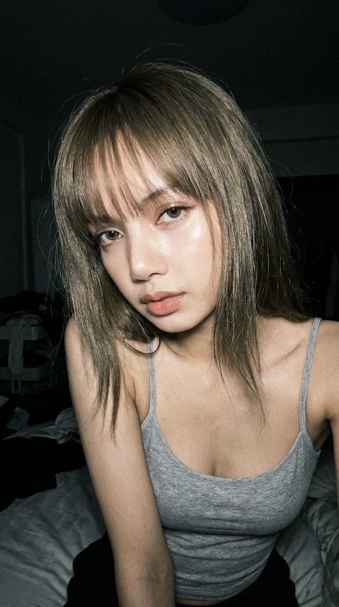 Portrait of a woman leaning toward the camera in a dim bedroom, showcasing a bold gaze with glossy skin reflections.