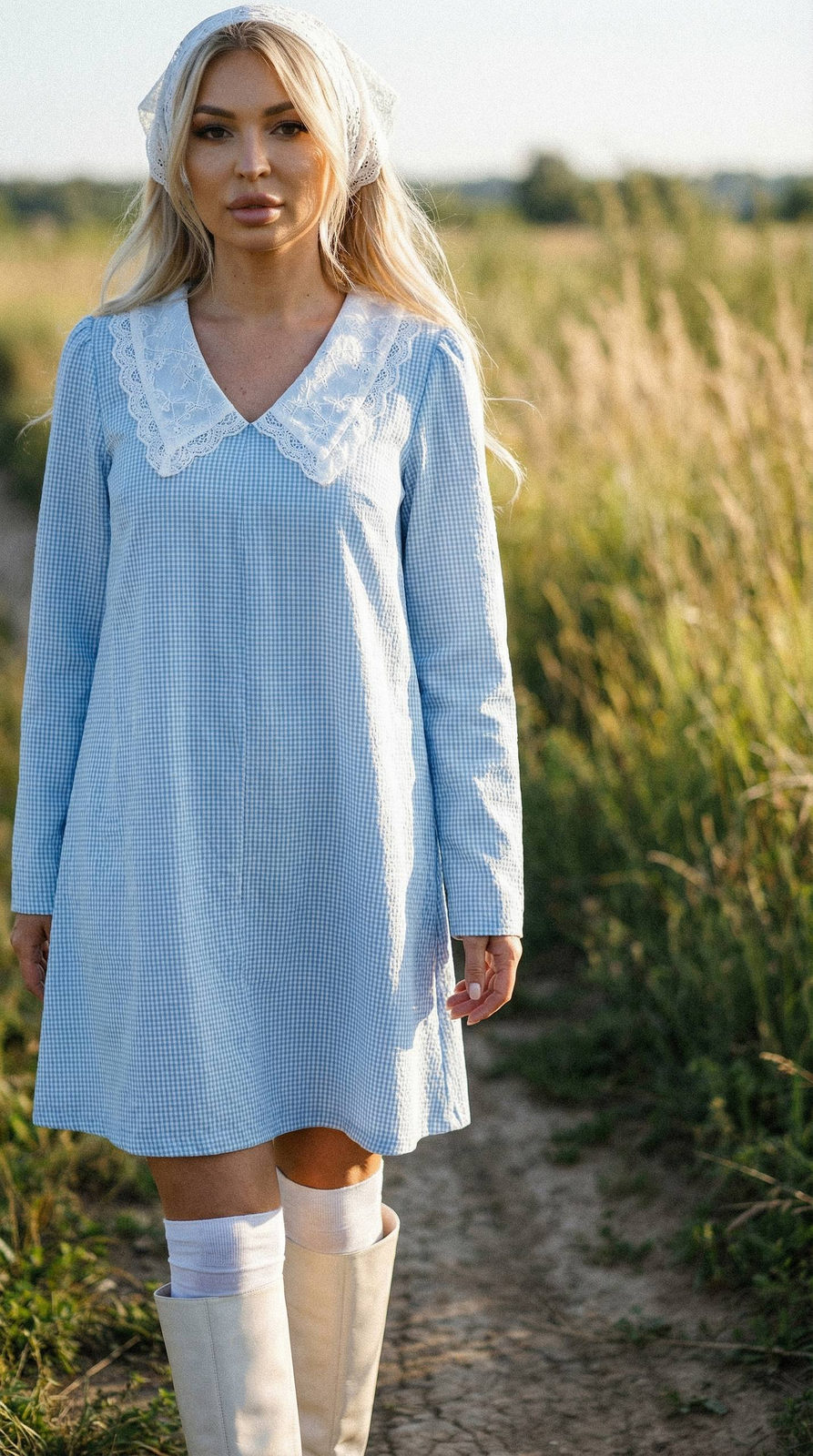 Portrait of a girl in a blue checkered dress and white lace headscarf, standing on a field path in daylight.