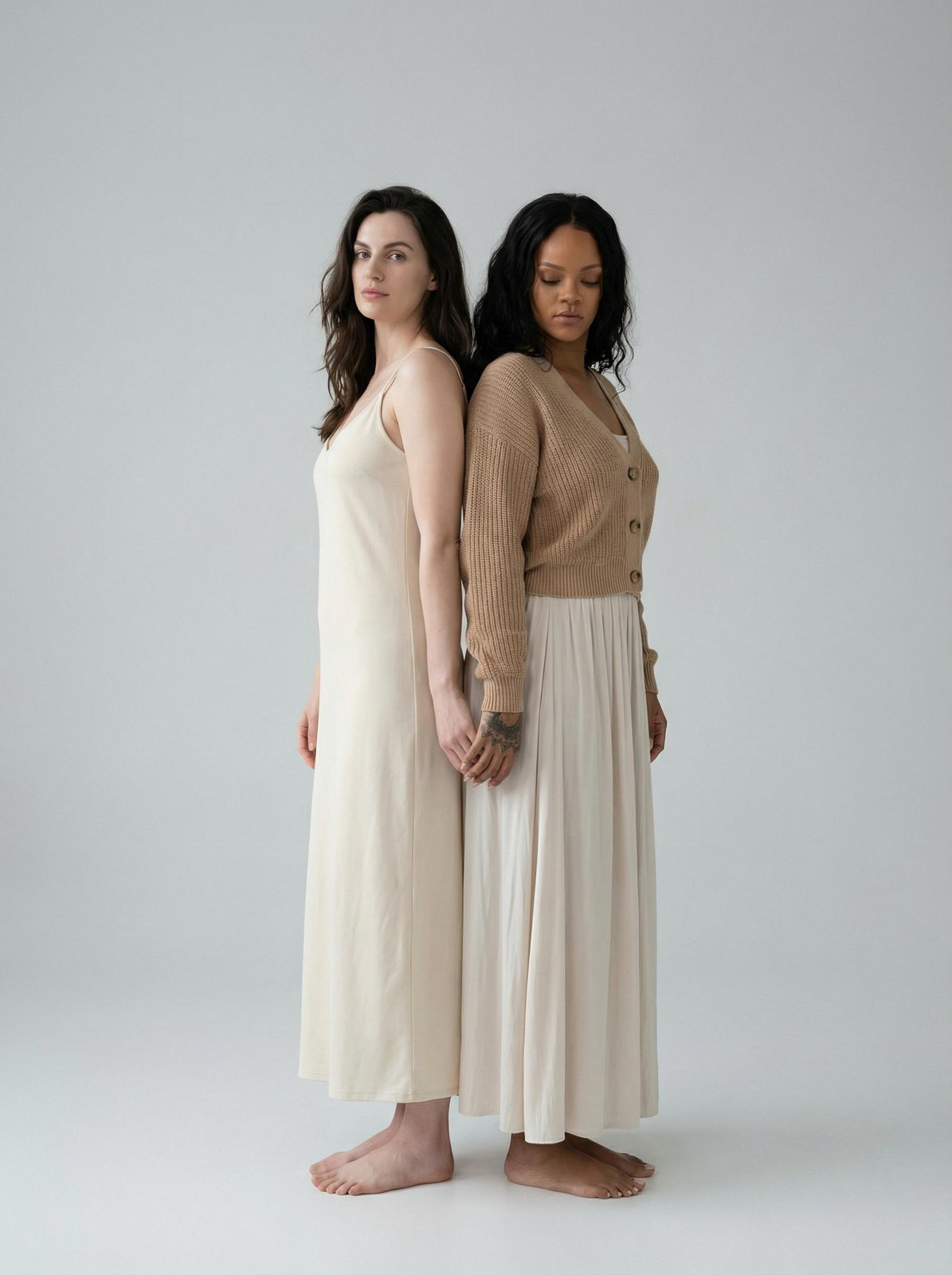 A full-figure portrait of two women standing back to back in a minimalist studio, softly lit with natural makeup.