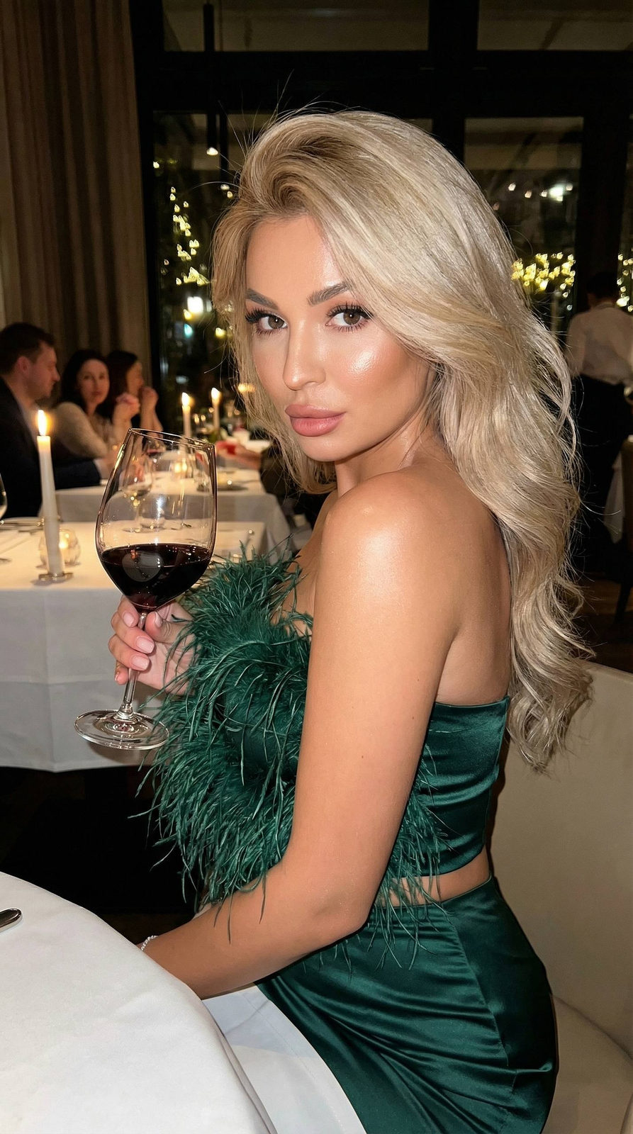 Beautiful girl in dark green silk outfit seated at a candlelit restaurant table, holding a glass of red wine, warm glowing lights in background.
