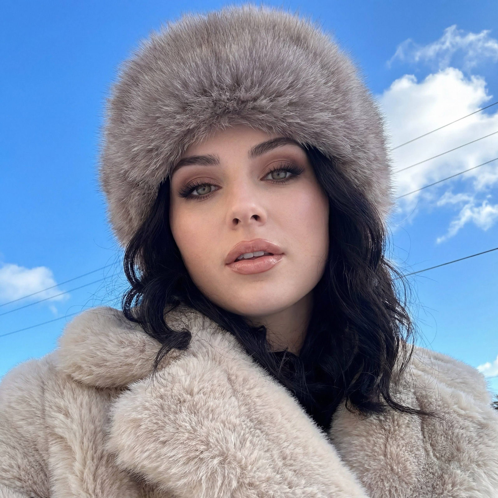 A close-up selfie of a confident woman in a fur coat, looking seductively at the camera against a bright blue sky.