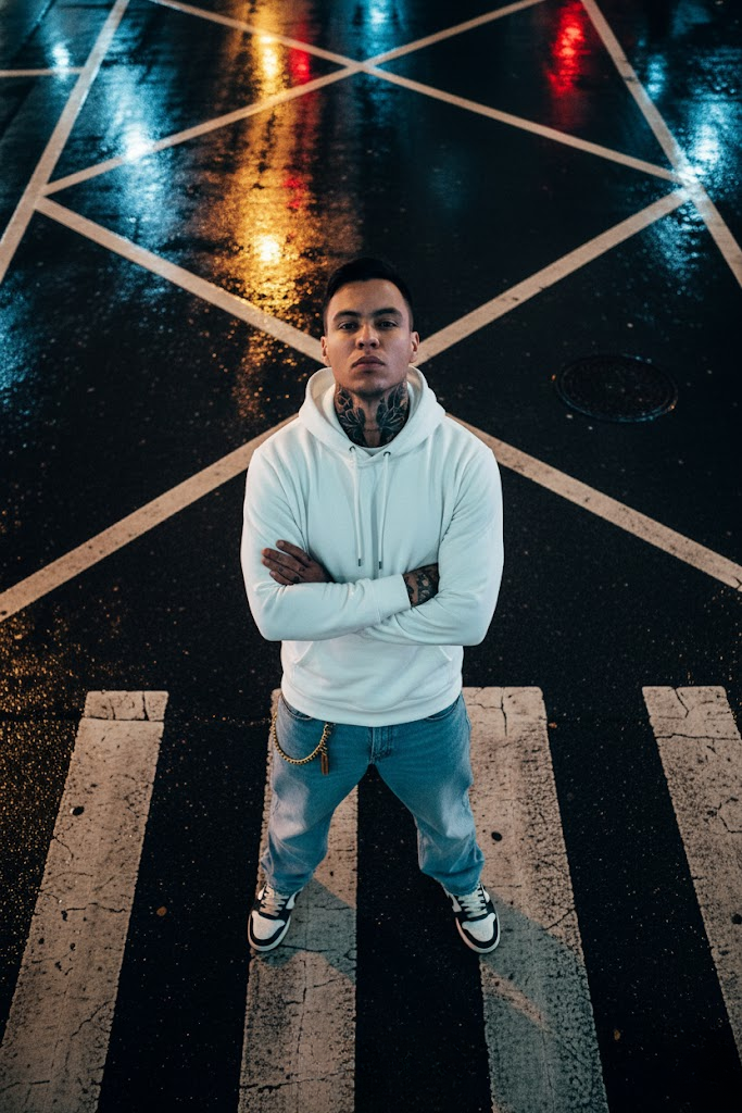 A striking streetwear portrait on a rain-slicked crosswalk, showcasing a person in a white hoodie with tattoos and sneakers, illuminated by streetlight reflections.