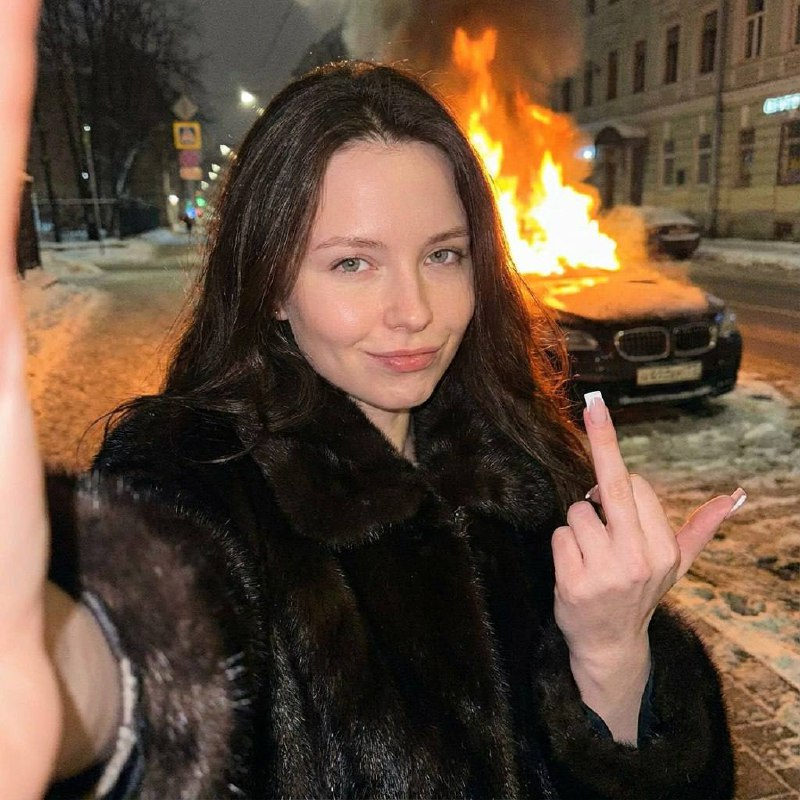 Close-up shot of a woman in a dark luxury fur coat smirking while showing her middle finger, with a burning luxury car in the background.