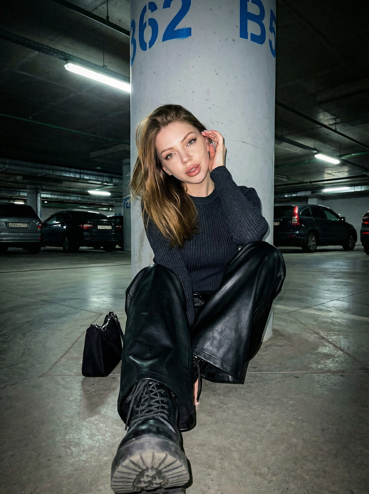 A woman with brown hair casually leaning against a concrete pillar in an underground parking lot, wearing a dark top and leather trousers.