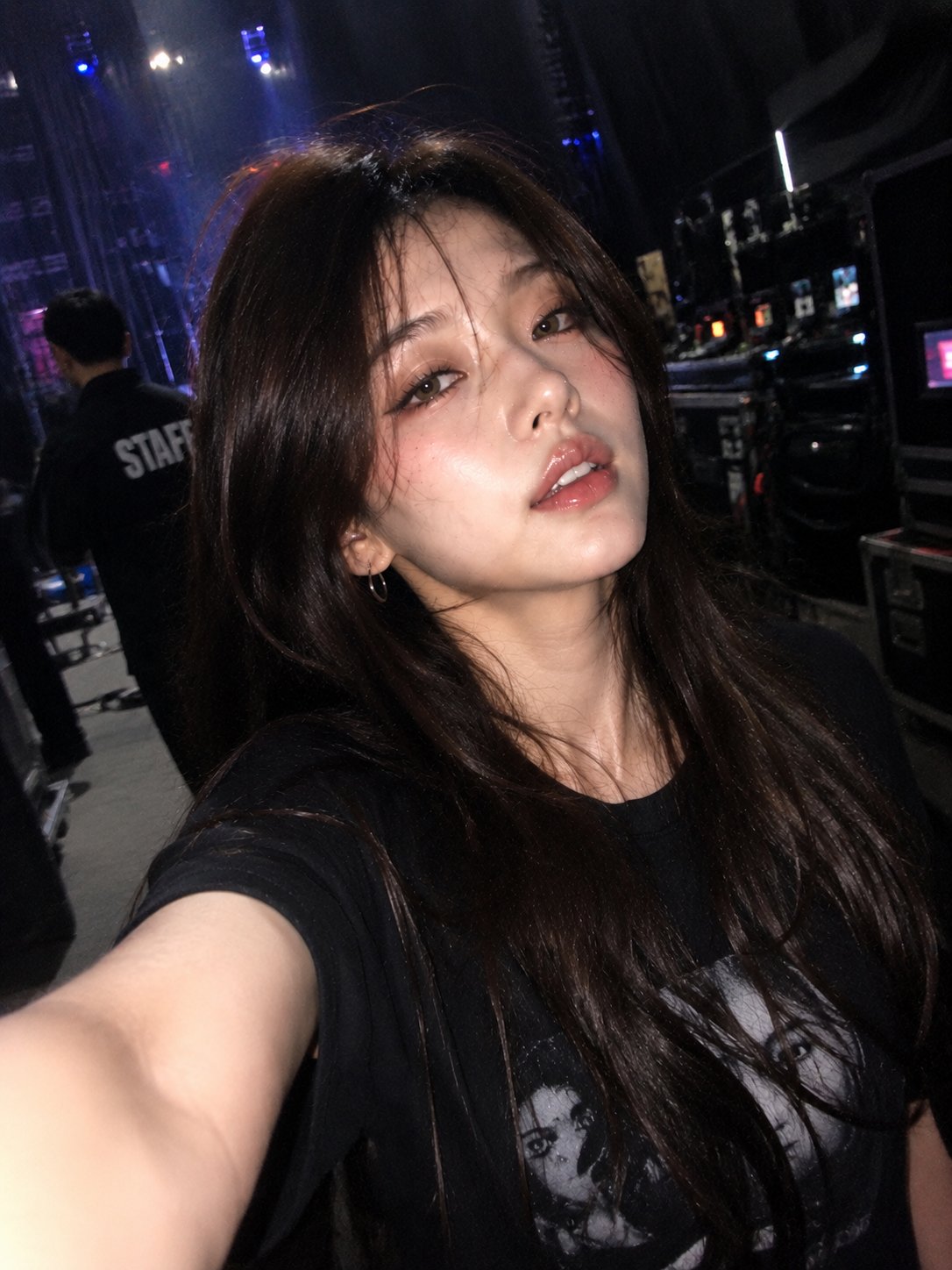 A high-angle photo of a person in a black graphic t-shirt with a faded print, backstage at a concert, illuminated by blue stage lights.