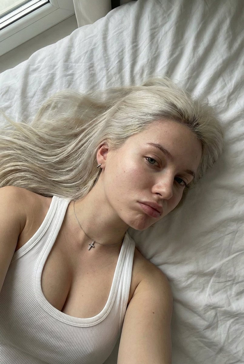 A young woman with platinum blonde hair lying on a bed, soft lighting accentuating her delicate features.