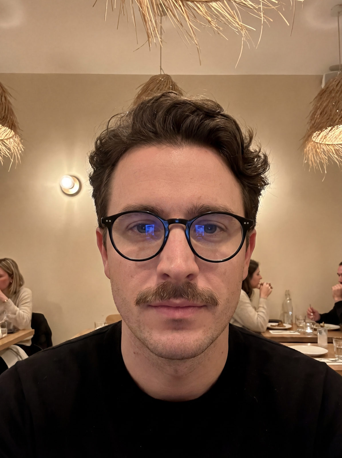 Close-up portrait of a man with round glasses and a defined mustache in warm indoor lighting.
