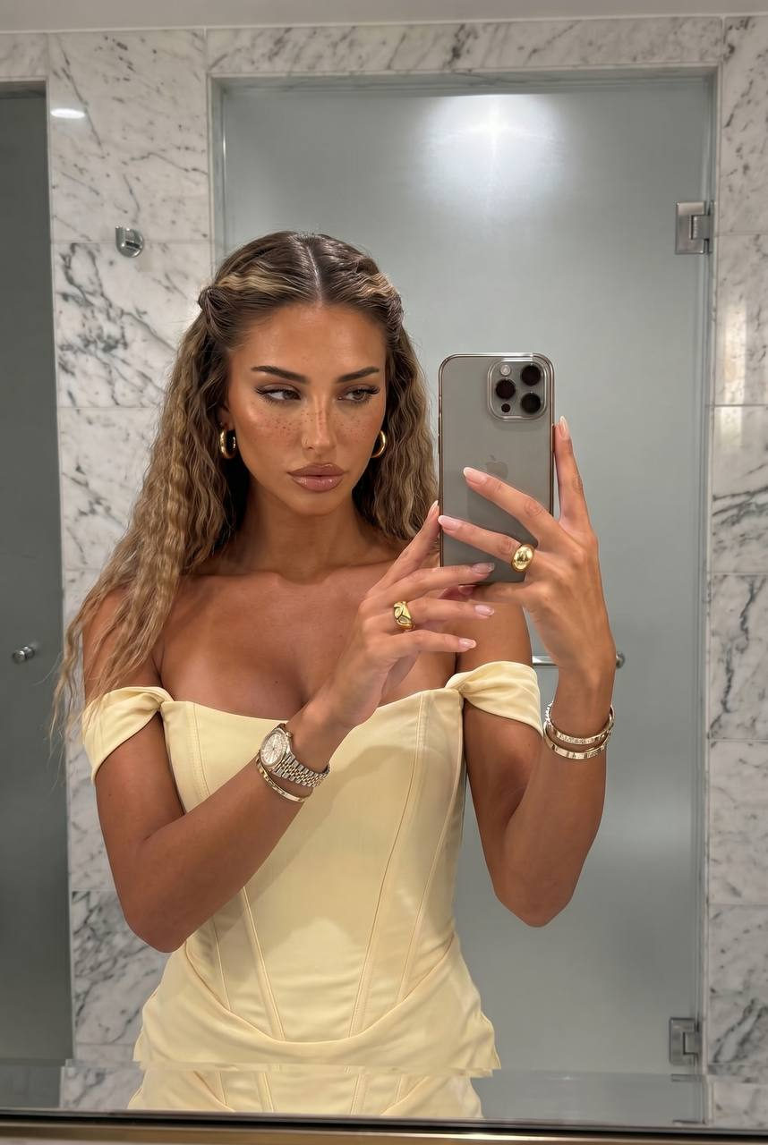 A woman in a pale yellow corset dress taking a mirror selfie in a marble bathroom with elegant lighting.
