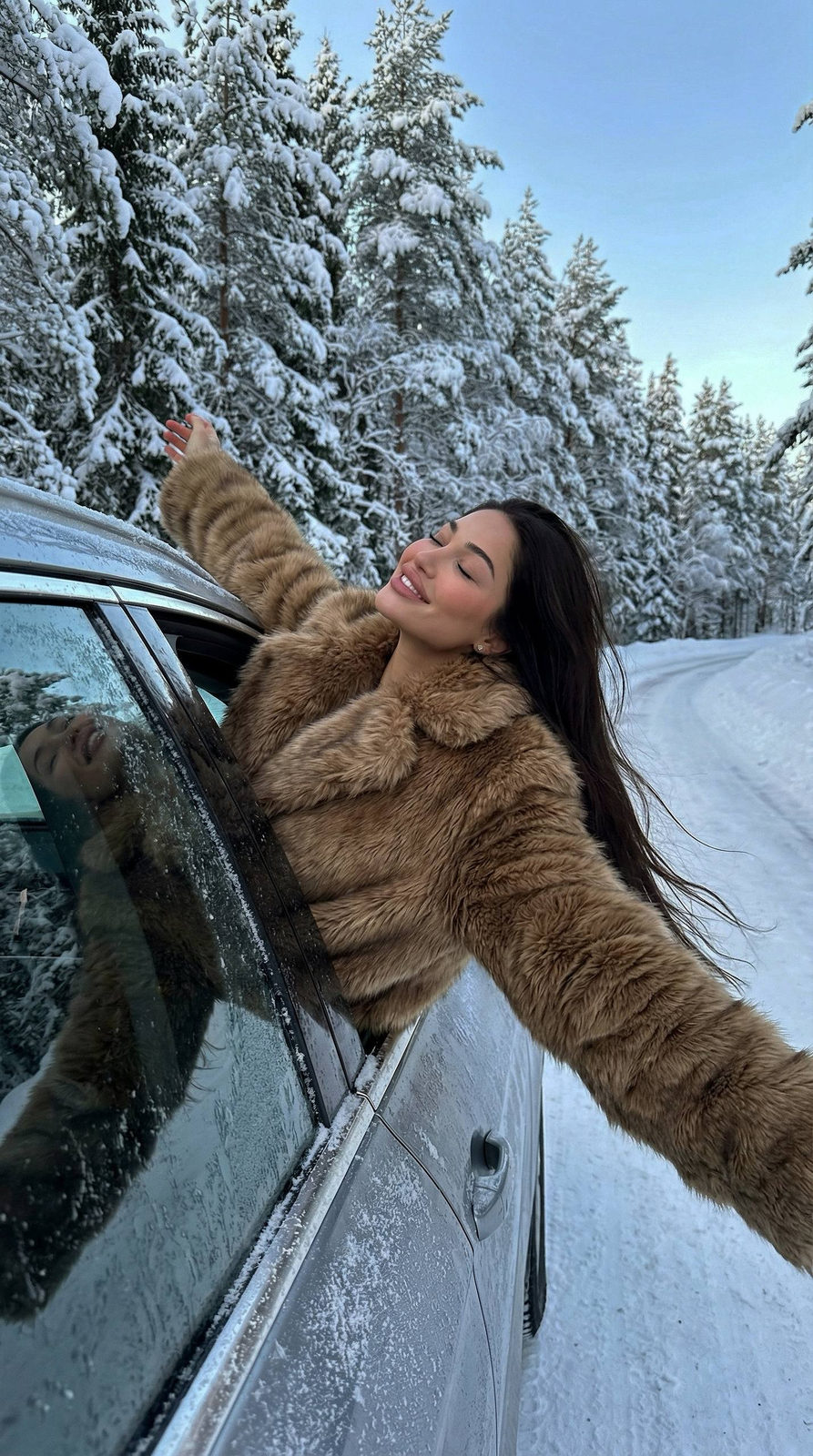 A joyful woman leans out of a moving car window on a snowy road, wearing a faux-fur coat, arms stretched wide against the cold air.