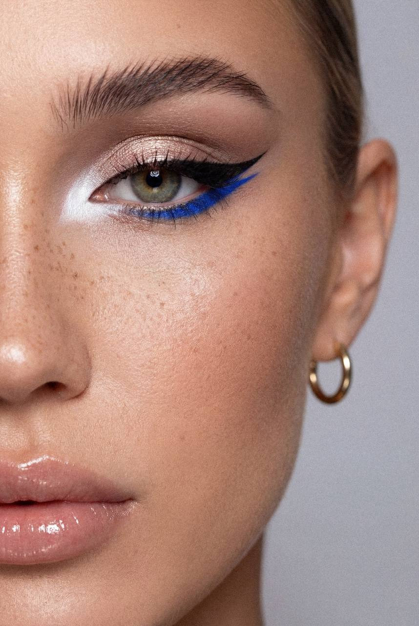 Ultra-realistic close-up of a female eye with bold graphic eyeliner and metallic champagne eyeshadow against a soft gray background.