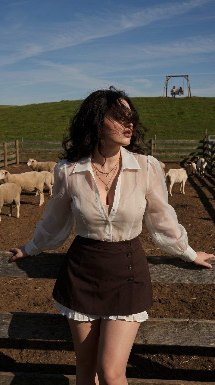 Stylish woman with dark wavy hair leaning on a wooden fence, wearing a semi-transparent blouse and A-line skirt, in a rustic farm setting.