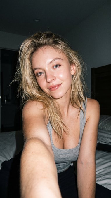 High-angle close-up selfie of a person in a heather gray tank top on a bed, featuring soft shadows and a blurred bedroom background.