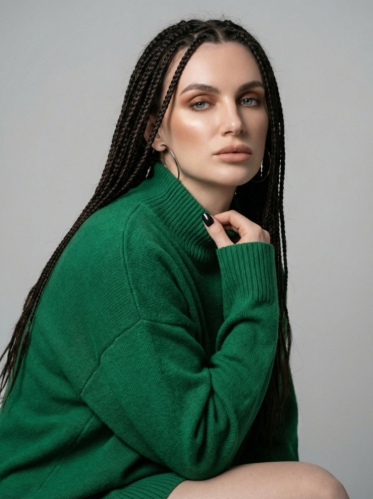 A woman in a green oversized sweater poses in a minimal studio. Her detailed braids and warm makeup stand out against a light gray background.