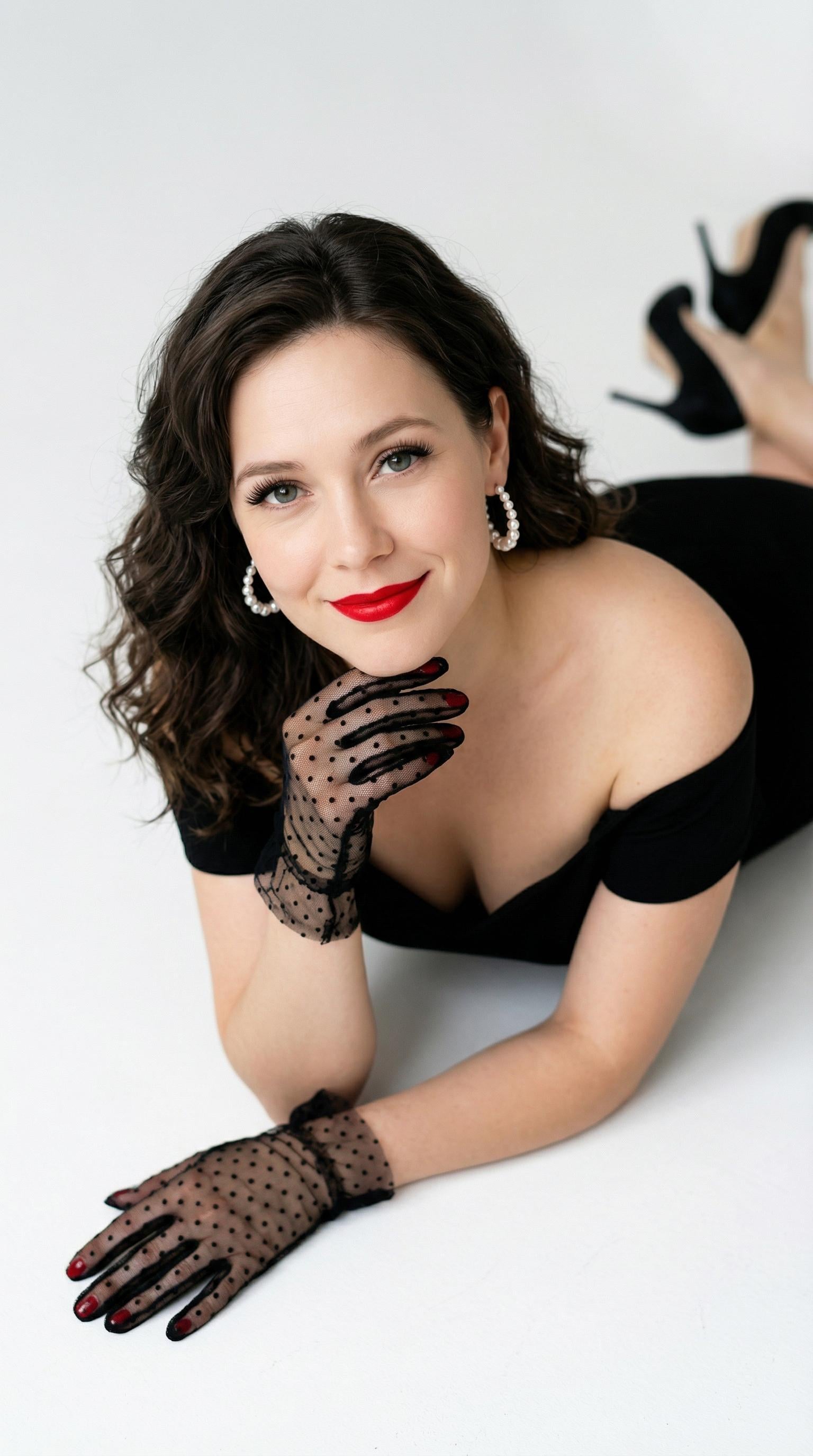 A woman in a black off-the-shoulder dress reclining on a clean white floor, wearing pearl hoop earrings and sheer black lace gloves