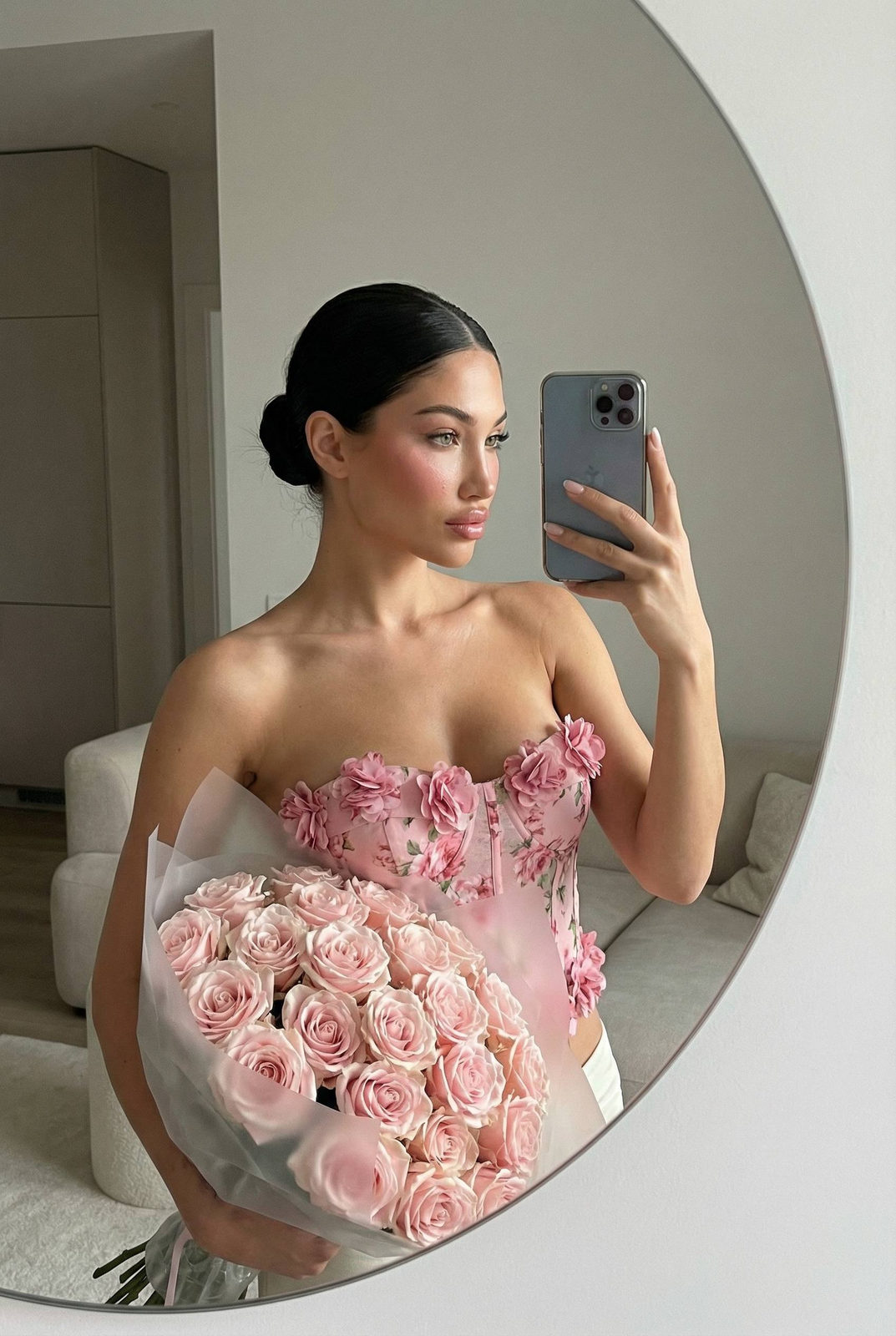 A woman in a floral corset top holds a bouquet of pastel pink roses in front of a rounded mirror, capturing her reflection.