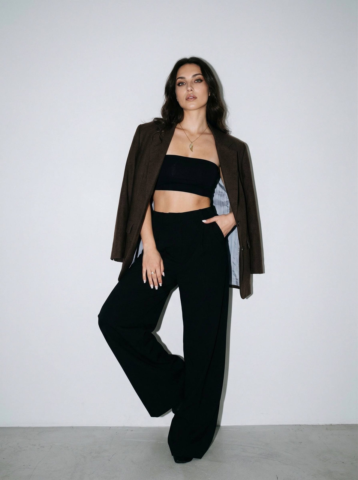 A model in a black strapless crop top with oversized blazer, showcasing a relaxed pose against a light gray background.