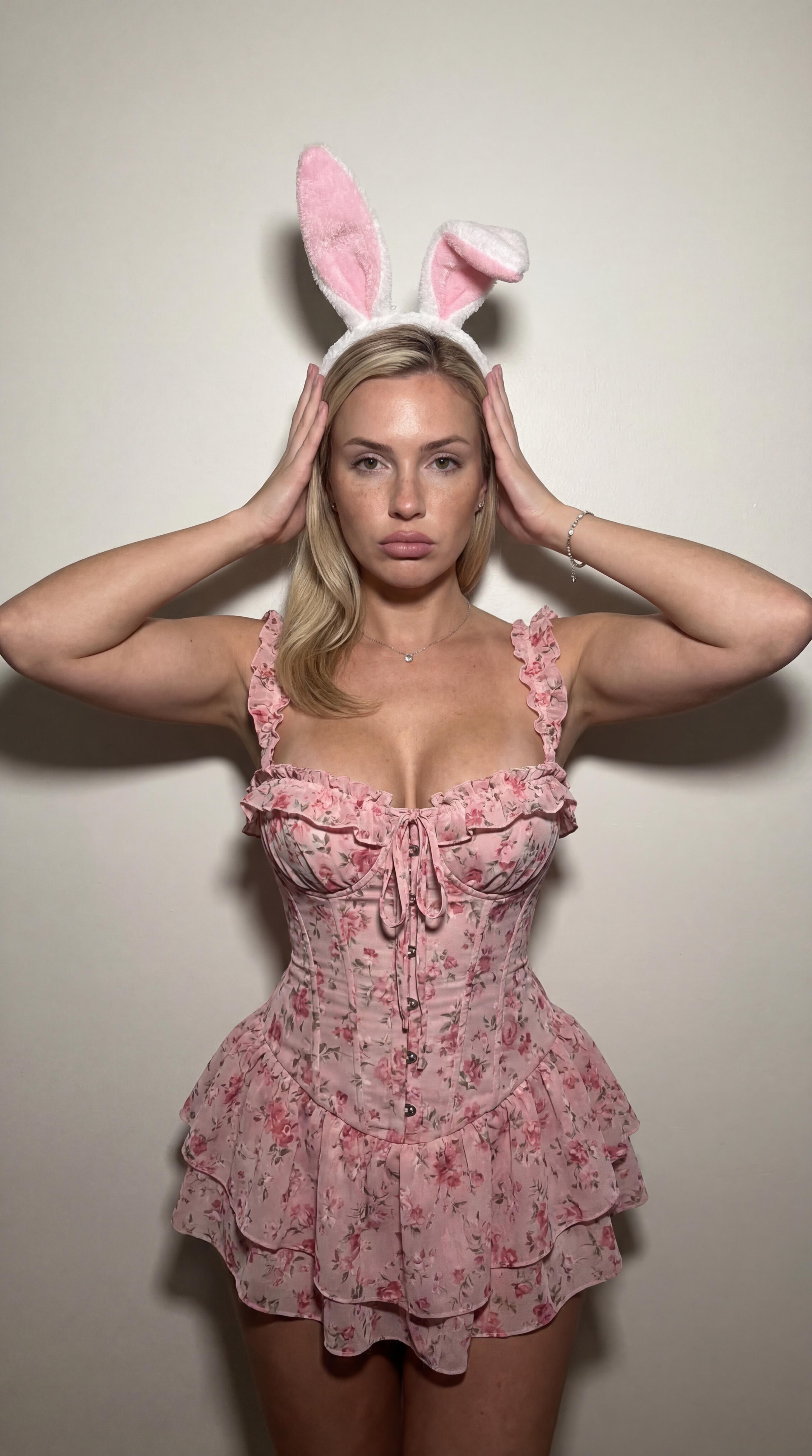 A person wearing a pink floral corset with ruffled trim and bunny ears, captured in soft diffused light against an off-white wall.