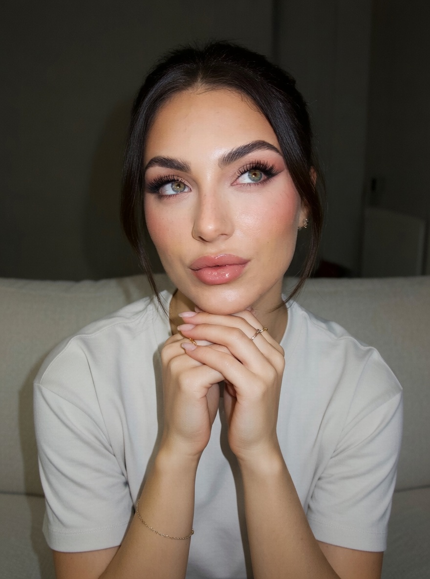 Close-up of a thoughtful woman sitting on a soft sofa, hands raised to her face with pastel eyeshadow and a soft pink gloss on her lips.