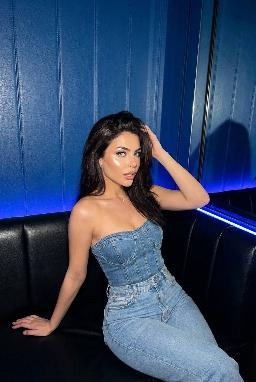 Ultra-realistic portrait of a woman in a denim corset on a leather sofa, looking playfully at the camera with bright makeup.