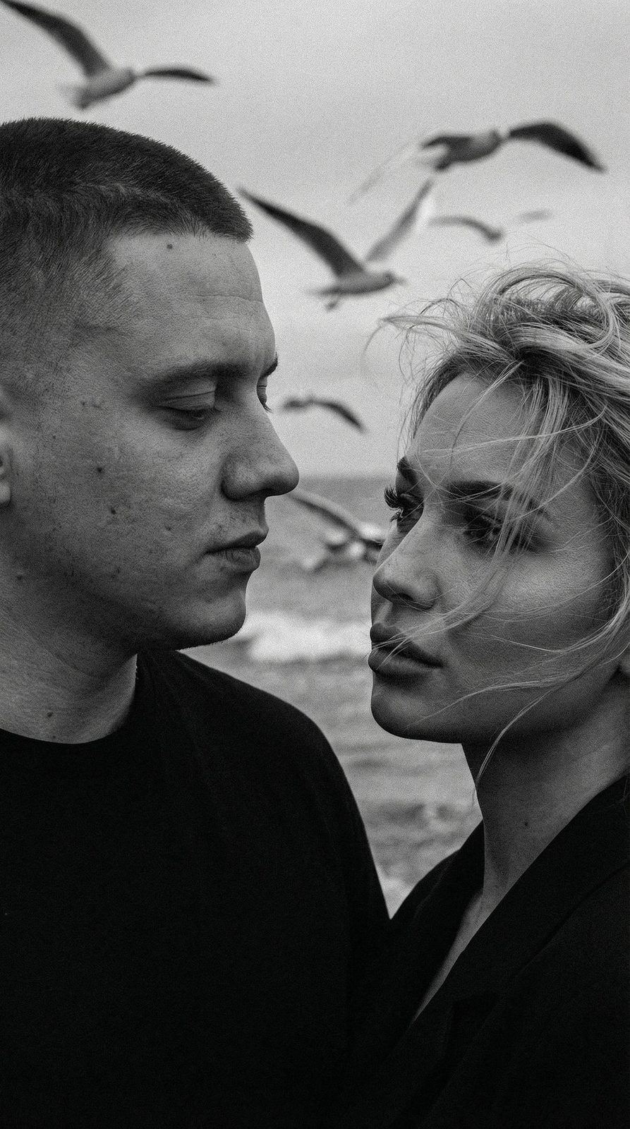 Close-up of a dramatic couple by the sea, wind ruffles hair with seagulls in the background.