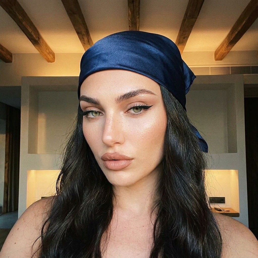Close-up selfie of a woman with a satin bandana, confident gaze, and flawless makeup under soft, warm lighting.