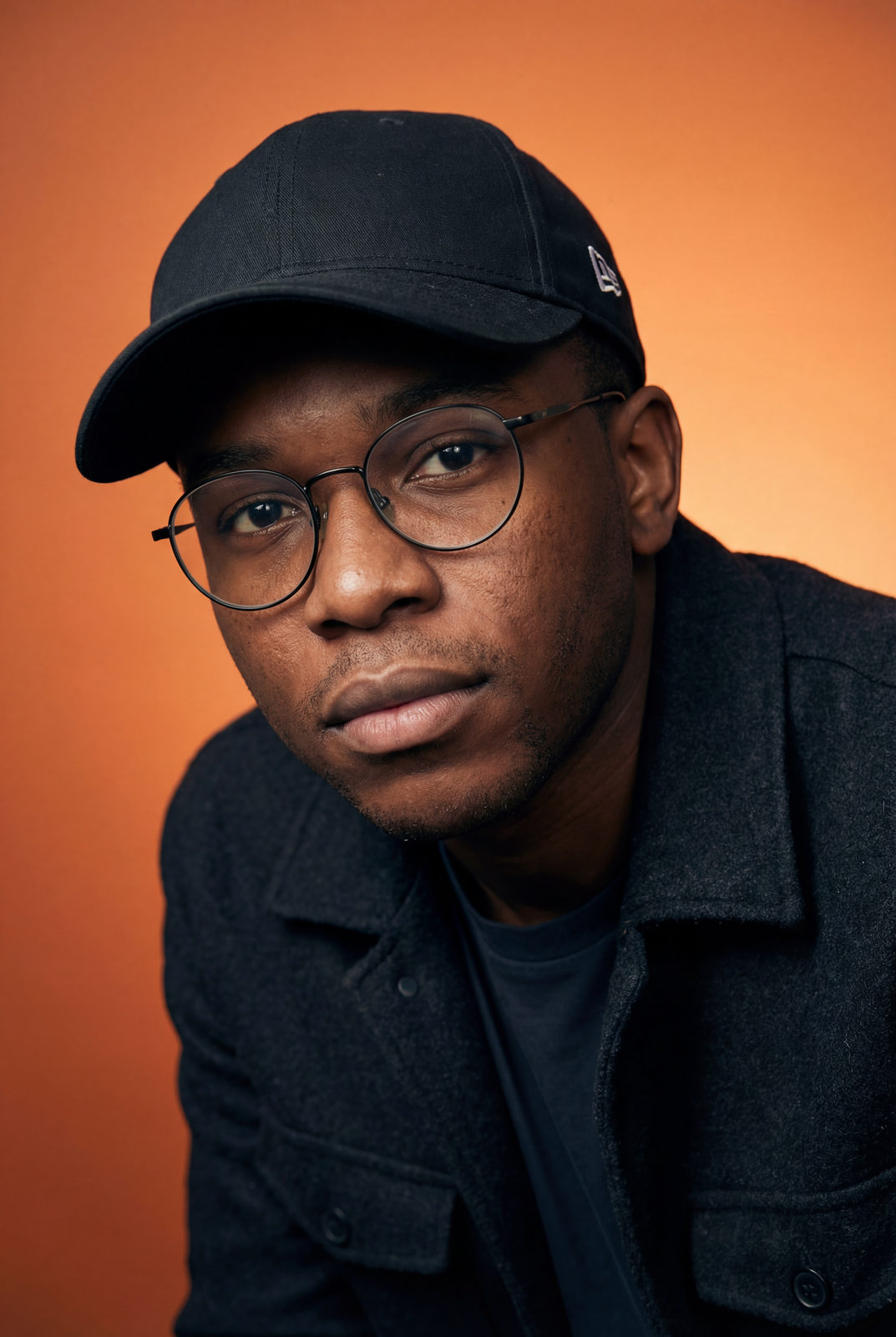 Ultra-realistic studio portrait of a person with a baseball cap and glasses, leaning forward against an orange gradient background.