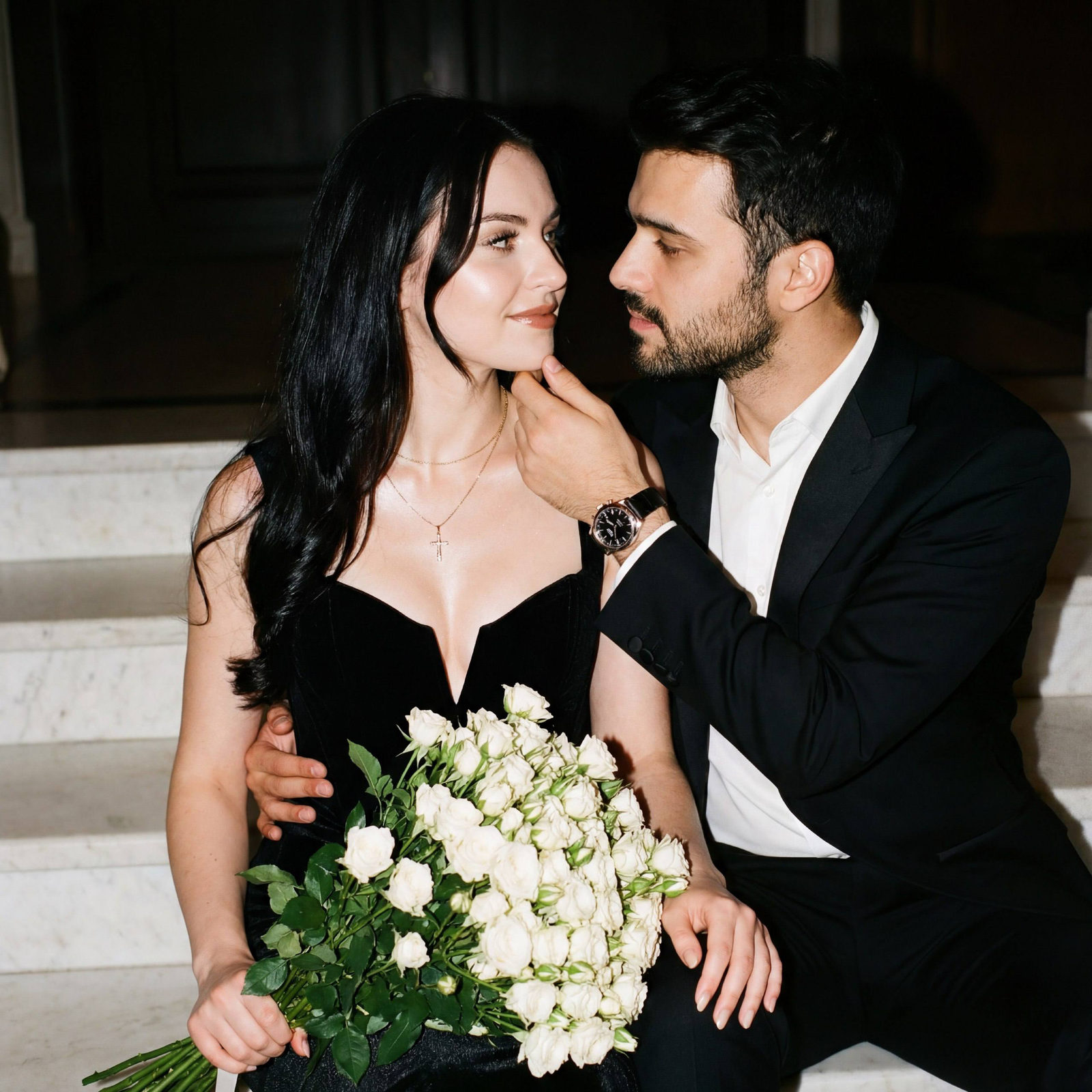 A woman in a black velvet corset holding a bouquet of white roses, sitting near a man in a tuxedo on a white marble staircase.