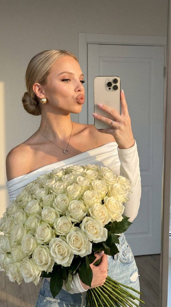 A woman with a playful pout in a large mirror, wearing a white top and jeans, holding a bouquet of white roses.