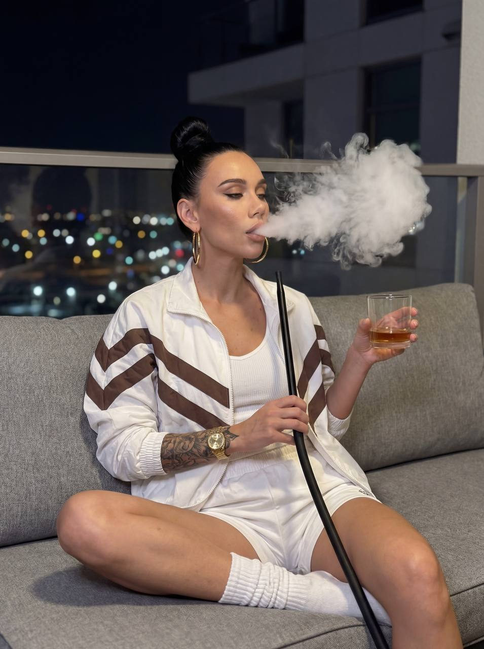 A woman with black hair in a high bun, exhaling smoke from a hookah while seated on a grey outdoor sofa at night.