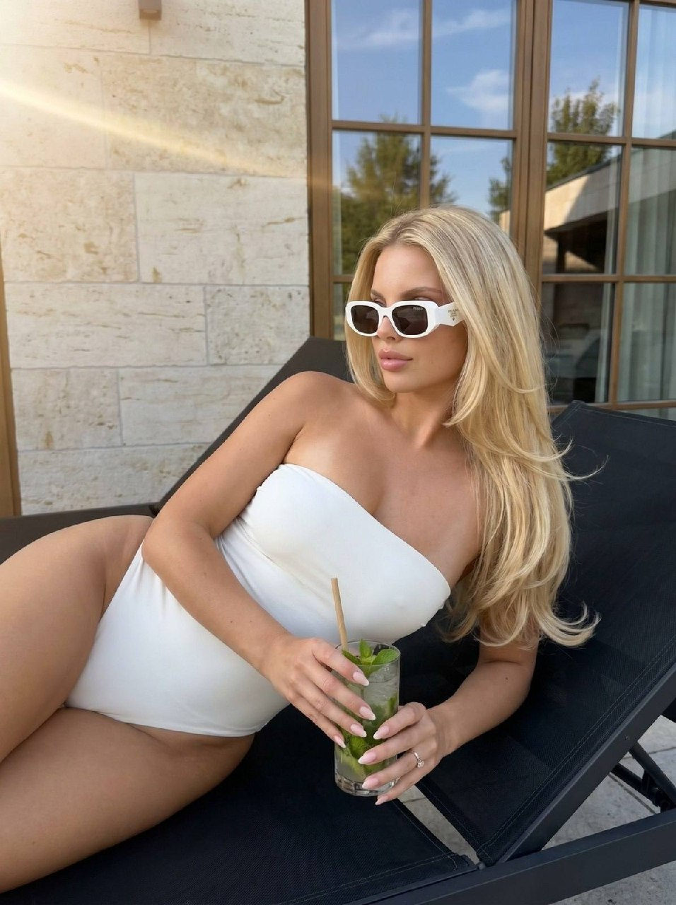 A slender tanned woman in a white swimsuit holding a mojito on a black sun lounger against a light stone wall.
