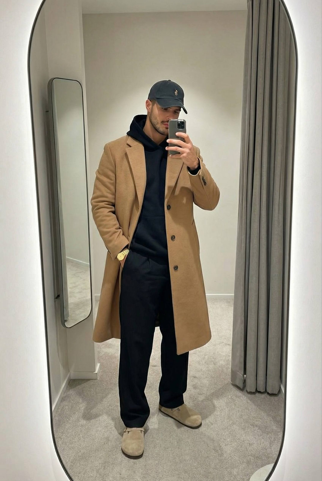 Realistic mirror selfie of a man in a fitting room, casually posed with a smartphone.