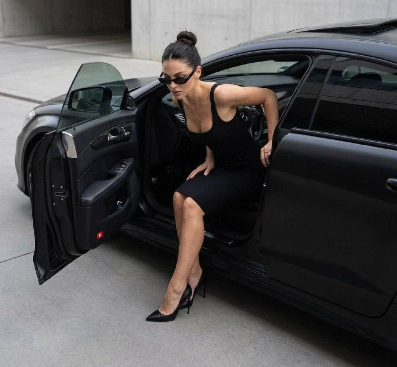 A graceful woman in stylish narrow dark glasses exiting a black matte Mercedes, showcasing her elegant posture and high stiletto pumps.