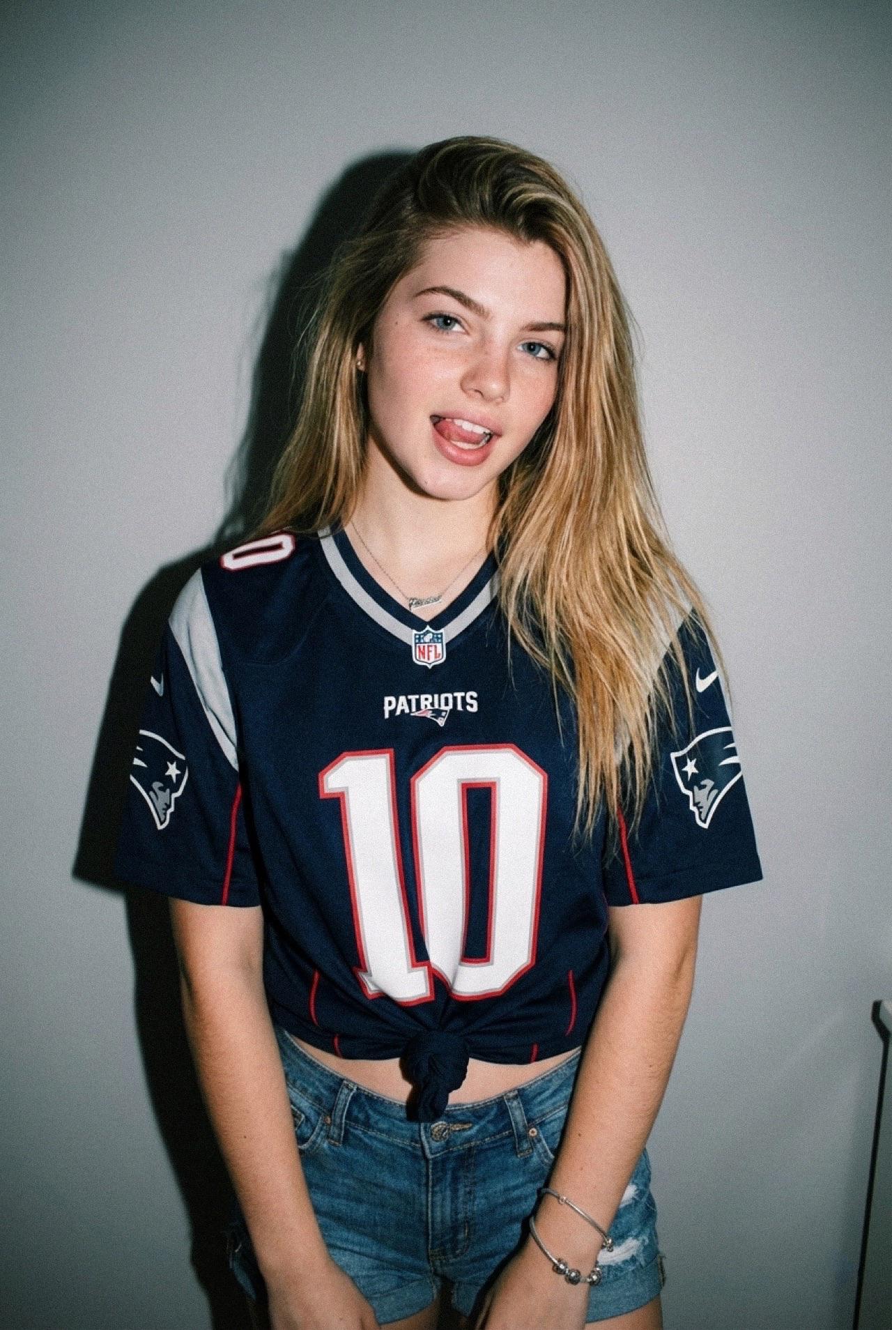 A playful portrait of a person wearing a navy blue Patriots jersey tied at the waist, revealing distressed denim shorts, tongue sticking out.