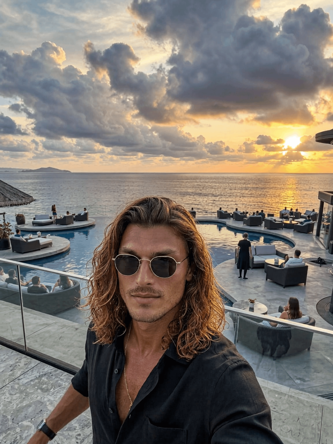 A person in round sunglasses and a black shirt relaxes by an infinity pool at sunset with dramatic clouds reflecting golden light.
