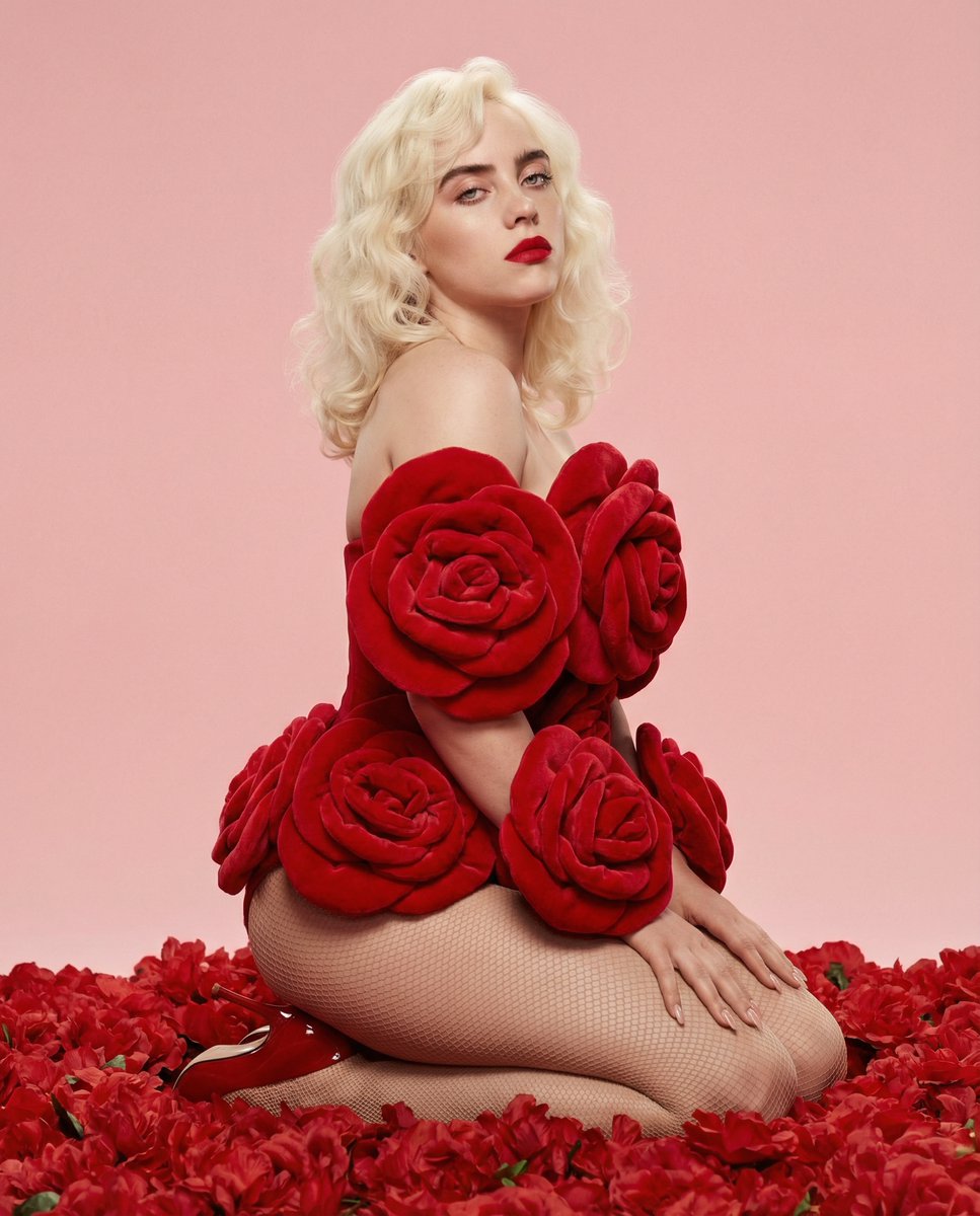 A medium-full body portrait of a model kneeling in a dense red floral arrangement, featuring soft pastel pink background.