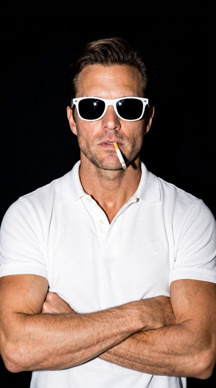 Close-up portrait of a man in a white polo and stylish glasses, holding a cigarette against a deep black background.
