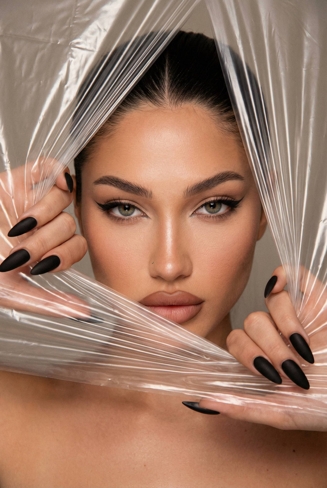 A close-up portrait of a female model behind translucent plastic film, showcasing elegant fingers and dramatic makeup.