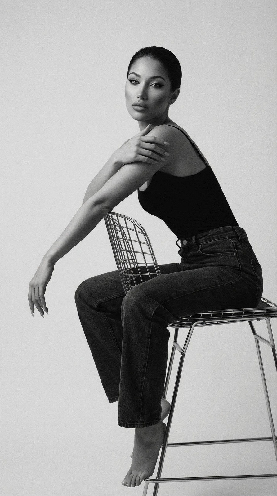 High-contrast studio portrait of a person on a metal wire chair, showcasing sculptural geometry and minimalist aesthetics.