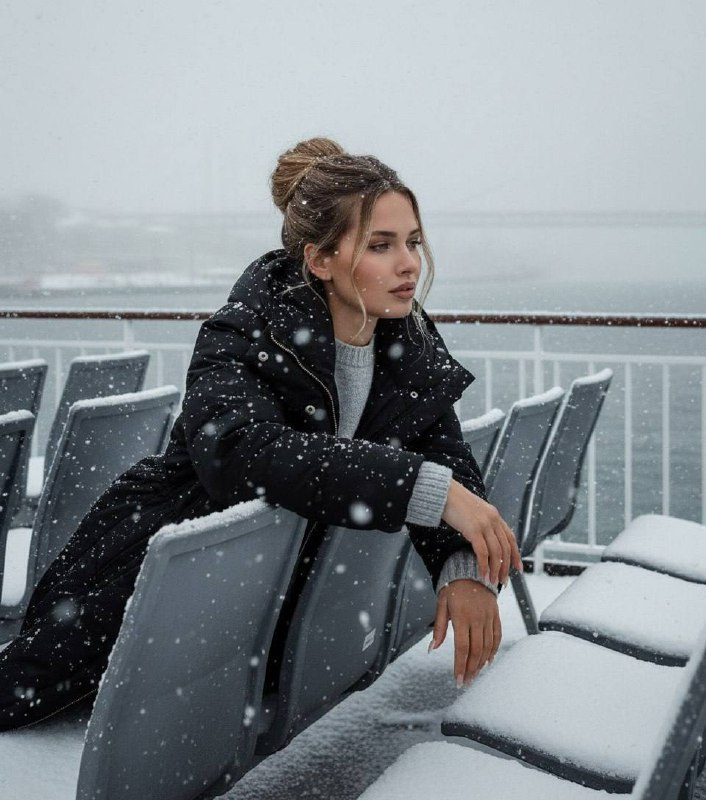 A thoughtful woman in a bulky winter coat leans against a snow-covered ferry seat during a heavy snowfall, with a blurred cityscape behind her.