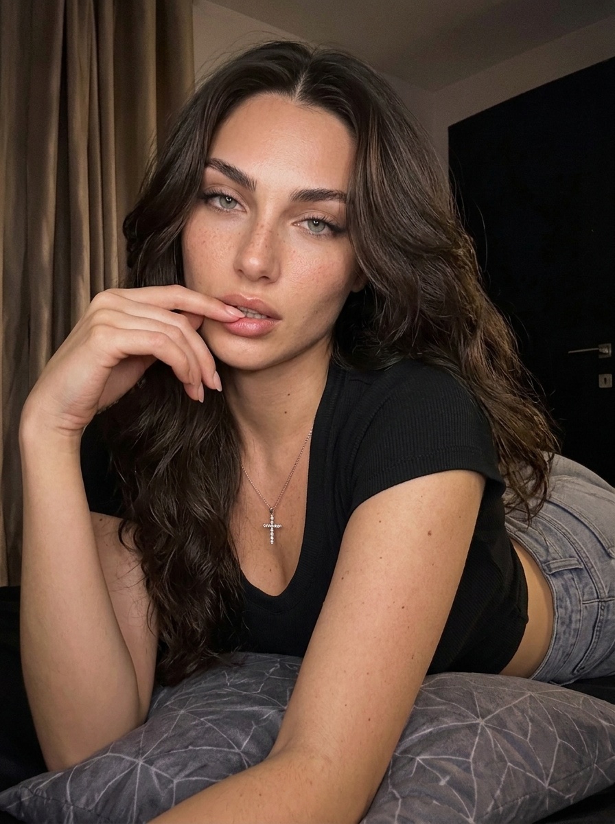 Lily leaning on a gray patterned pillow, wearing a black cropped top and denim shorts, in dim light with soft shadows on her face.