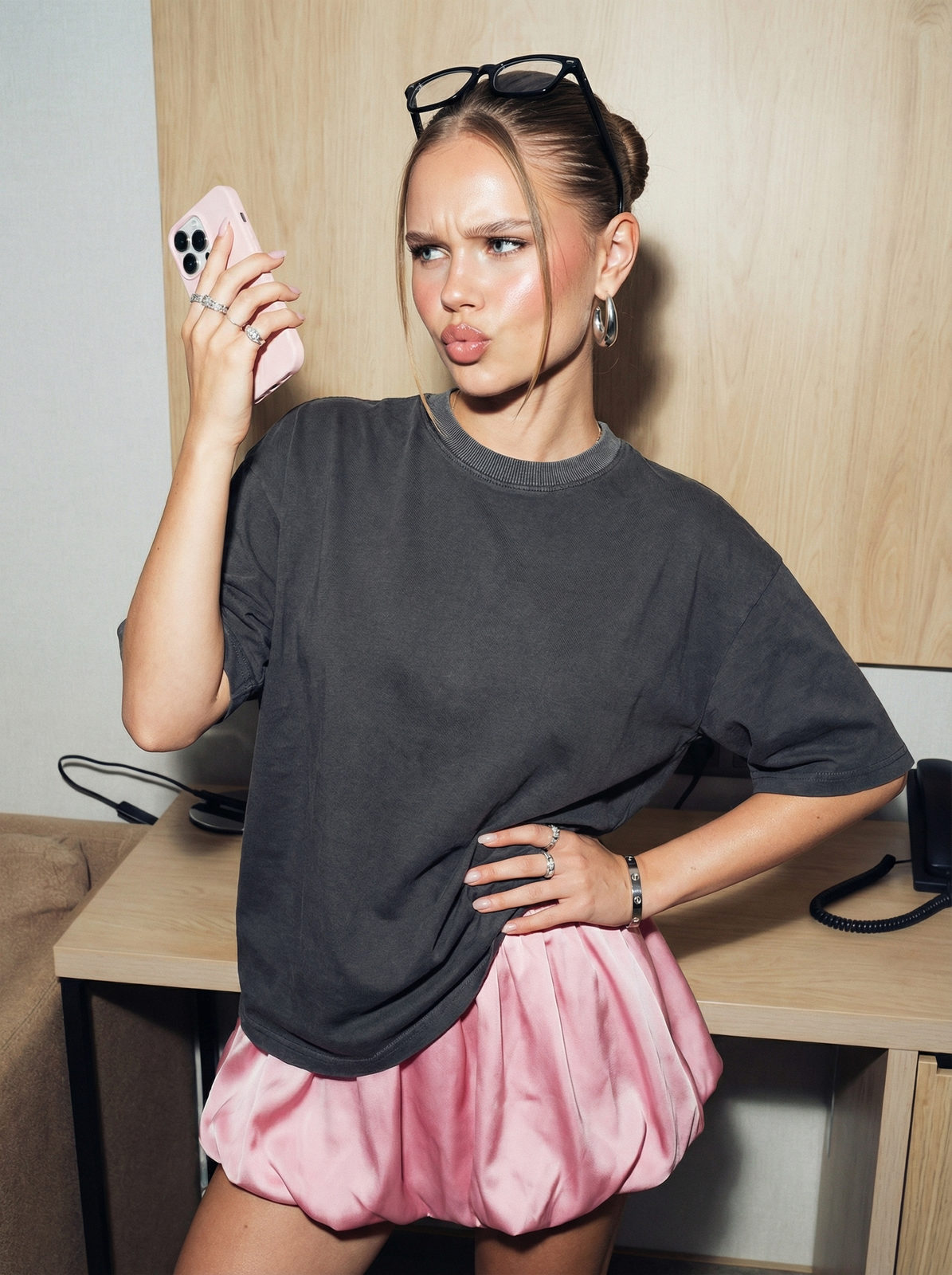A young woman in a grey oversized cotton t-shirt and pink silk balloon skirt, posed elegantly near a modern hotel table.