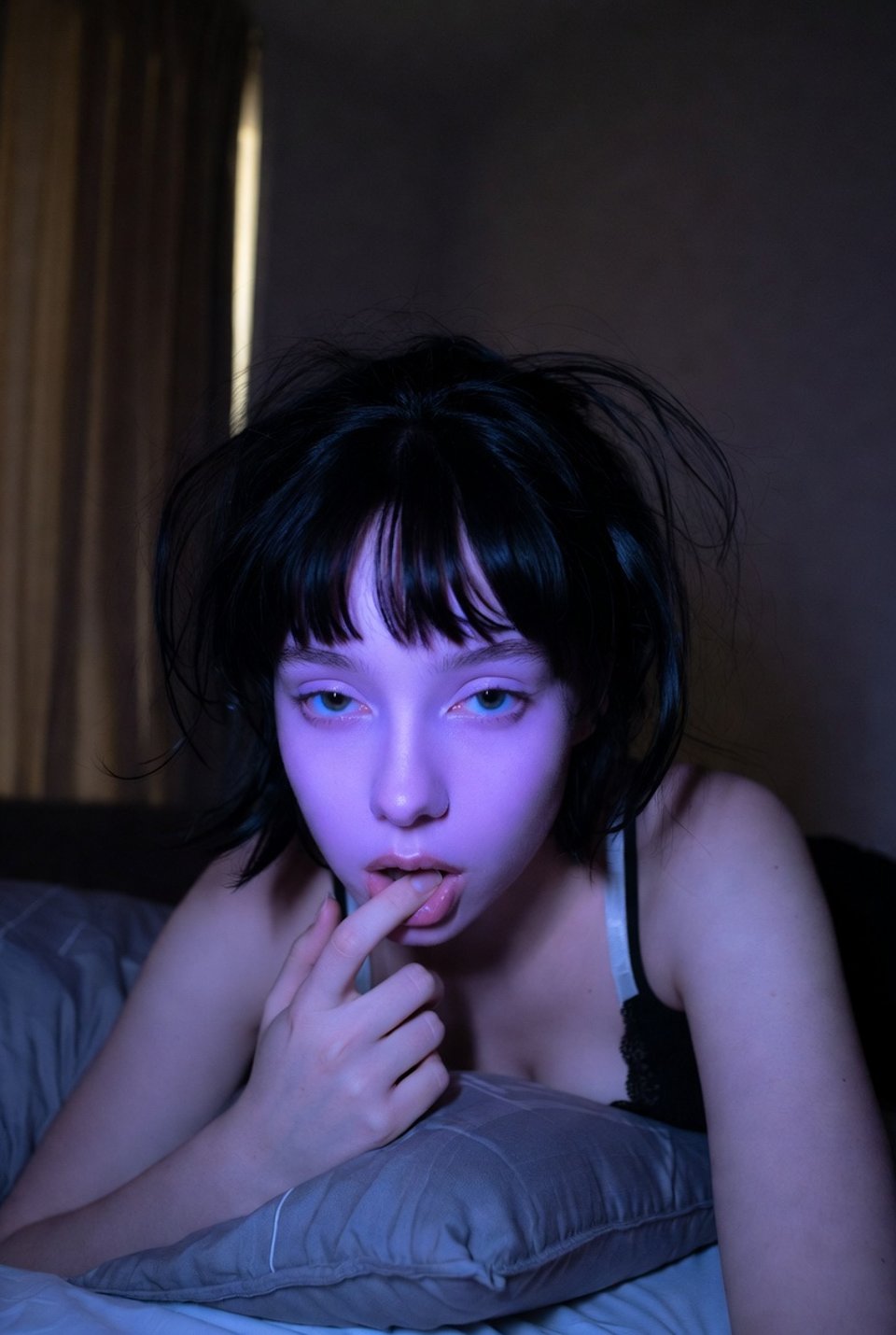 A low-light phone selfie in a dimly lit bedroom, showing a pale face illuminated by bluish-purple screen glow.