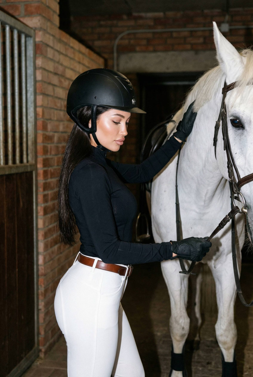 A woman in a fitted black riding top stands beside a white horse in an equestrian stable, showcasing a calm expression and interaction.